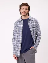 Camisa de manga larga con estampado de cuadros en tonos blanco, gris y azul. Presenta cuello clásico, cierre frontal con botones y un pequeño parche con logo en el pecho.