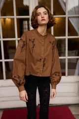 Camisa de algodón color blanco con cuello mao y cierre frontal de botones. Presenta un diseño de bordados florales en color negro distribuidos en el frente y las mangas. Posee mangas largas abullonadas con puños abotonados y un corte recto ligeramente cropped.