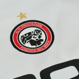 Camiseta de fútbol blanca con detalles en rojo y negro. Tiene cuello redondo y mangas cortas. En el frente se lee la palabra "PALCO" en letras negras y el logo de Palace en el pecho.