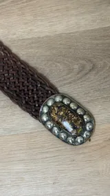 Cinto trenzado de cuero color chocolate con hebilla ovalada de metal envejecido, decorada con una piedra central jaspeada y tachas metálicas alrededor.