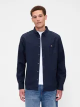 Camisa Oxford de manga larga en color azul marino, con cuello abotonado, cierre frontal de botones y un bolsillo en el pecho que presenta un pequeño logo bordado de un oso.