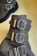 Bota de caña media confeccionada en cuero color gris oscuro con efecto desgastado. Presenta tres correas laterales con hebillas metálicas grandes y una suela track negra de plataforma dentada.