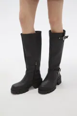 Bota de caña alta color negro, confeccionada en eco cuero, con suela track de goma y detalle de hebilla metálica en el tobillo y en la parte superior de la caña.