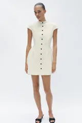Vestido corto blanco de encaje, con cuello redondo, manga corta y bajo evasé con frunces. Cuenta con fila de falsos botones negros delanteros a contraste y cierre trasero con cremallera oculta.