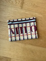 Clutch de neopreno negro con cierre superior. Presenta un estampado de cuadros en tonos beige, marrón y azul, con letras grandes en color vino tinto que forman la palabra "MILO" y pequeños dibujos temáticos.