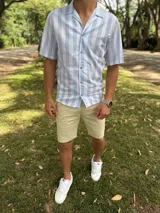 Camisa de manga corta con diseño a rayas verticales en color celeste y blanco, cuello abierto estilo cubano y cierre frontal con botones marrón claro.