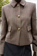 Blazer corto de corte estructurado con cuello solapa, manga larga y hombreras. Presenta cierre frontal con botones metálicos y dos bolsillos delanteros de plastrón con solapa. Diseño con estampado de cuadros tipo príncipe de gales.