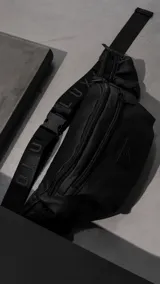 Riñonera negra confeccionada en cordura impermeable de alta resistencia. Presenta tres compartimentos principales con cierres metálicos, un bolsillo lateral adicional y una correa ajustable de alta densidad con broche de seguridad. Incluye una chapa metálica decorativa en el frente.