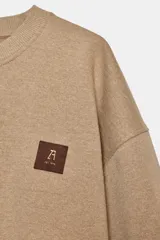 Sudadera de algodón color beige, con cuello redondo, manga larga y corte ligeramente corto. Presenta un pequeño parche cuadrado marrón con un logo grabado en el pecho.