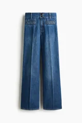 Pantalón de jean estilo wide leg con tiro alto, confeccionado en denim de algodón rígido. Presenta bolsillos delanteros con detalle de ribete trenzado, pliegues frontales y traseros, y cierre con doble botón.