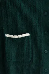 Saco corto de punto trenzado fino en color verde oscuro, con cuello redondo, cierre frontal de botones y bolsillos delanteros. Presenta detalles de ribetes festoneados en color crema en los bolsillos y en los puños.
