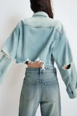 Cazadora corta de denim en tono celeste claro, con cuello solapa, manga larga y cierre frontal con botones. Presenta un bolsillo de plastrón en el delantero y detalles de rotos en el cuello y el ruedo deshilachado.