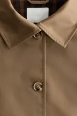 Chaqueta corta de sarga color marrón tostado, con cuello clásico, mangas raglán y cierre frontal con botones. Cuenta con bolsillos laterales, cordón ajustable en el bajo y puños con vuelta que dejan ver un forro a cuadros.