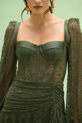 Vestido corto color verde oliva, con cuerpo tipo corset de escote recto y mangas largas de encaje. La falda presenta un diseño drapeado con volados laterales y textura de encaje.