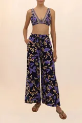 Pantalón de corte amplio con estampado de hojas y manchas en tonos violeta y beige sobre fondo negro. Cuenta con aberturas laterales y cintura elástica ajustable con cordón.