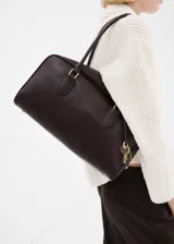 Bolso de mano de poliuretano color marrón oscuro, con doble asa para el hombro, herrajes metálicos dorados y detalle de llavero decorativo con candado y llaves.