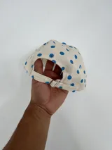 Gorro tipo jockey color beige con lunares azules, visera azul y bordado frontal con la palabra "ABOIU" en letras de colores y estrellas.