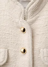 Chaqueta de tweed color crema con cuello mao, cierre frontal de botones dorados efecto joya y cuatro bolsillos delanteros con solapa. Presenta diseño recto, manga larga y forro interior.