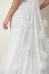 Vestido de novia blanco strapless con corsé armado y falda corte sirena de tul drapeado. Presenta apliques de flores de organza en la falda y una cola romántica de tul.