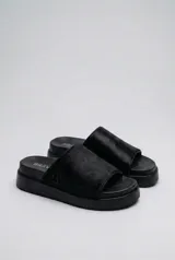 Sandalias planas tipo mule negras, con tira ancha superior de pelo y suela de caucho expandido. Llevan el logo BRAVA bordado en el lateral de la plantilla.