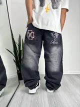 Pantalón de jean estilo baggy con efecto lavado desgastado en color negro. Presenta bolsillos traseros con bordados distintivos: uno con un diseño geométrico en rojo y blanco, y otro con una figura angelical y la inscripción Ameri.