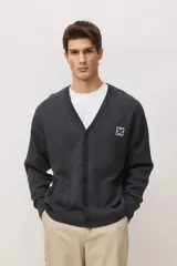 Cardigan de punto en color gris oscuro melange, con cuello en V, cierre frontal con botones y bolsillos delanteros. Presenta un parche cuadrado con una X en el pecho.