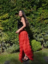 Falda larga de lino color rojo, con aberturas laterales y múltiples niveles de volados formados por flecos de rafia. Cuenta con cintura elástica en la parte posterior.