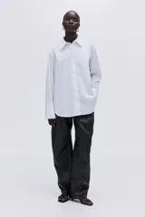 Camisa blanca de popelina de algodón, corte oversized con cuello clásico, hombros caídos y mangas largas con puños anchos abotonados. Presenta cierre frontal de botones y bajo redondeado.