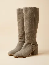 Botas de caña alta desmontables color visón, confeccionadas en gamuza natural. Presentan un diseño de punta semi cuadrada, taco ancho de 6 cm y detalle de estribo con hebilla forrada en la base.