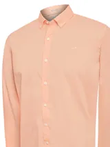 Camisa de manga larga color durazno, confeccionada en popelina de algodón orgánico. Tiene cuello camisero, cierre con botones y logo bordado en el pecho.