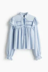 Blusa celeste de manga larga con textura arrugada, cuello alto con volados y abertura frontal con lazos para atar. Presenta canesú con volados y detalles de encaje calado en el pecho y hombros. Los puños terminan con elástico y volado.