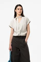 Camisa de calce relajado color beige claro, con escote en V y lazo largo incorporado para anudar al cuello. Cuenta con mangas cortas con vuelta, frunce sutil en los hombros y cierre frontal con botones.