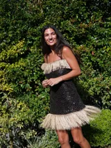 Vestido corto sin mangas, color negro, completamente cubierto de lentejuelas brillantes. Presenta un borde de flecos largos de color rosado en el escote y en el ruedo inferior.