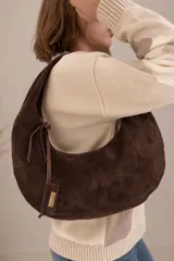 Cartera tipo morral confeccionada en gamuza color marrón chocolate, con diseño hobo de líneas curvas, asa integrada al hombro y detalles de tiras de cuero con placa metálica grabada.