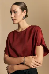 Top de manga corta color bordó, de silueta amplia y caída fluida, confeccionado en un tejido liviano con leve brillo. Presenta detalle de botón de nácar en la espalda con pequeña abertura y mangas amplias tipo kimono.