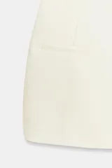 Mono corto color blanco de cuello subido y manga corta con hombreras. Presenta falsos bolsillos de vivo en el delantero y cierre frontal con cremallera y botón ocultos por sopata.