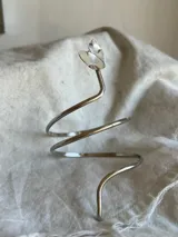 Brazalete rígido plateado con forma de espiral y detalle de hojas en uno de los extremos.