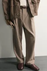 Pantalón relaxed fit de hombre color marrón con textura estructurada, cintura con pliegues frontales, bolsillos laterales y cierre frontal con cremallera y botón.