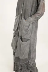 Camisa larga de silueta amplia en color gris, con cuello mao y cierre frontal de botones. Presenta hombros caídos, mangas largas semitransparentes, cuatro bolsillos delanteros grandes, ruedo redondeado y pliegue central en la espalda.