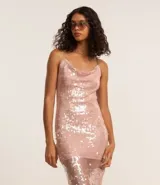 Vestido midi de lentejuelas color rosado, con escote drapeado en la parte delantera y escote degagê en la espalda con breteles finos cruzados. El bajo es asimétrico con punta y tiene un forro corto que crea un efecto de transparencia en las piernas.