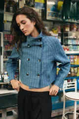 Chaqueta corta de cuello subido y manga larga, con diseño de cuadros en tonos azules. Presenta cierre frontal cruzado con botones oscuros y bolsillos de vivo en el delantero.
