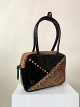Cartera rígida negra con detalles en rojo y tachas plateadas. Tiene herrajes y cierre metálico plateado marca YKK, asa fina para llevar al hombro o en la mano y correa larga extra regulable. El interior es forrado, tiene un bolsillo con cierre enmarcado en cuero, y un segundo bolsillo plaqué.