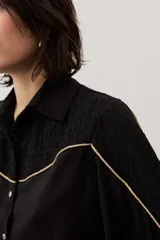 Camisa de manga larga color negro, con cuello camisero, cierre frontal con botones y detalles de ribetes contrastantes en color dorado. Presenta paneles superiores con textura y mangas abullonadas.