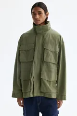 Campera estilo cargo de tejido ripstop, color verde militar, con cuello elevado que incluye capucha oculta. Presenta cierre frontal con solapa rompevientos, cuatro bolsillos delanteros con solapa y botón a presión, puños ajustables y cordón interno en la cintura.
