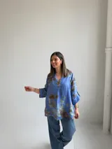 Blusa azul de lino con estampado floral en tonos marrones y verdes, escote en V con botón decorativo grande y mangas largas holgadas con puños arremangables.