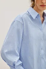 Camisa de corte recto y largo cropped, confeccionada en algodón con estampado de rayas verticales finas en color azul y blanco. Presenta cuello clásico, cierre frontal con botones y mangas largas con puños abotonados. La prenda está decorada con pequeñas aplicaciones de strass distribuidas por toda la superficie.