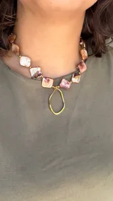 Collar corto con piezas cuadradas de nácar en tonos marrones y blancos, separadas por pequeñas cuentas doradas. Incluye un colgante central dorado con forma irregular.