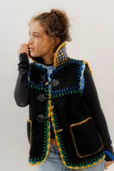 Chaleco negro de corderito con cierre de alamares, tejido a mano en crochet con detalles en amarillo, verde y azul. Cuello alto con interior a cuadros. Talle oversized.