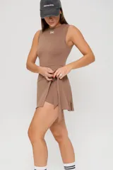 Vestido corto deportivo color habano, con diseño sin mangas, cuello redondo y detalle de falda envolvente con lazo lateral. Incluye logo pequeño bordado en el pecho.