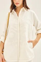 Campera de gabardina color crudo con cuello camisero, mangas largas regulables con tira y botón, y cierre frontal con botones ocultos. Presenta parches bordados decorativos en el delantero y la espalda, además de bolsillos laterales tipo ojal.